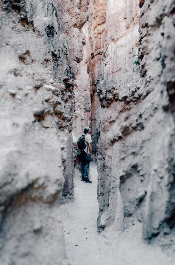 A hiker with a backpack navigates through a narrow, deep corridor between towering clay formations, highlighting the scale and the intricate textures of the eroded walls in Los Estoraques Unique Natural Area.