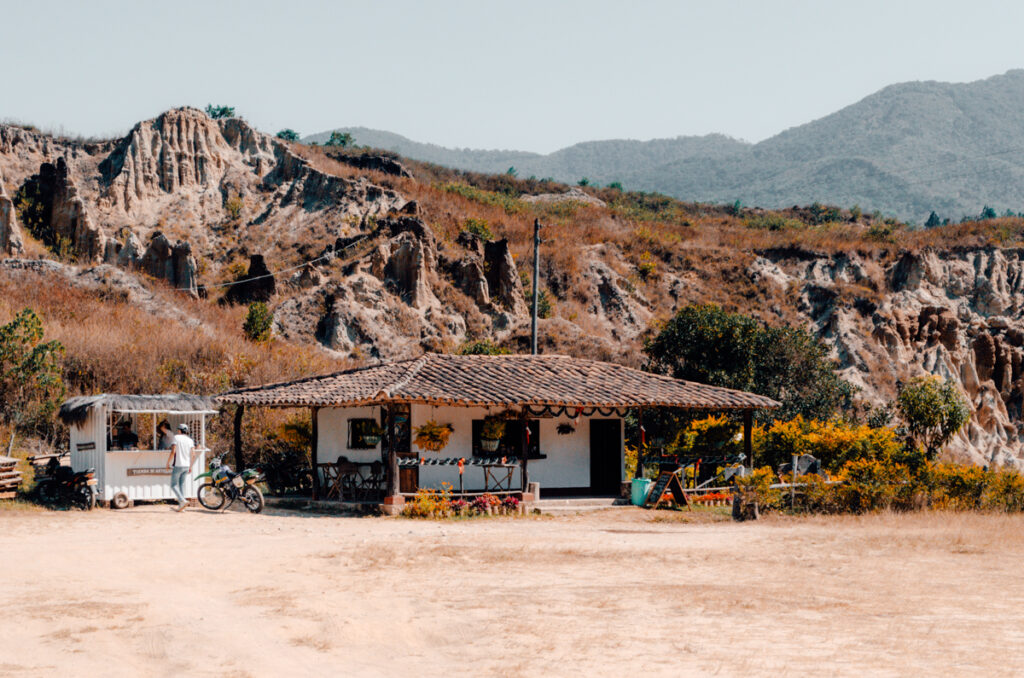A rustic countryside building with a thatched roof and a motorcycle parked out front, set against the dramatic backdrop of the eroded hills of Los Estoraques Unique Natural Area. This is where the entrance fee is collected and you can also get drinks and snacks.