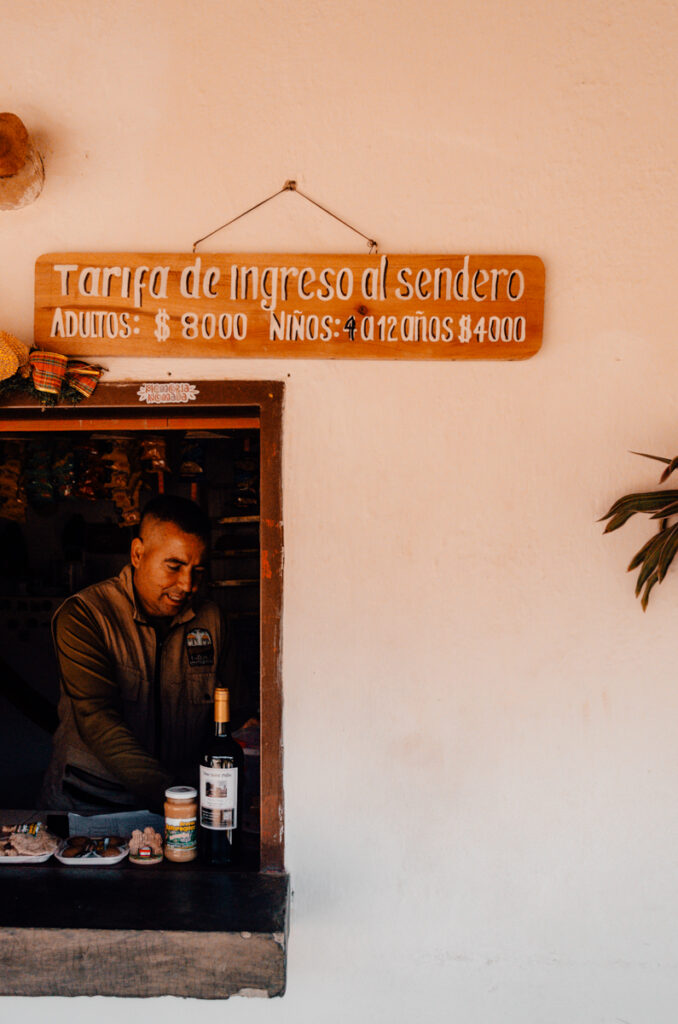 A trail entrance sign reading 'Tarifa de Ingreso al sendero' with prices listed for adults and children, hanging above a small kiosk window where a man is serving local food and drinks and collect entrance fees for Los Estoraques Unique Natural Area.