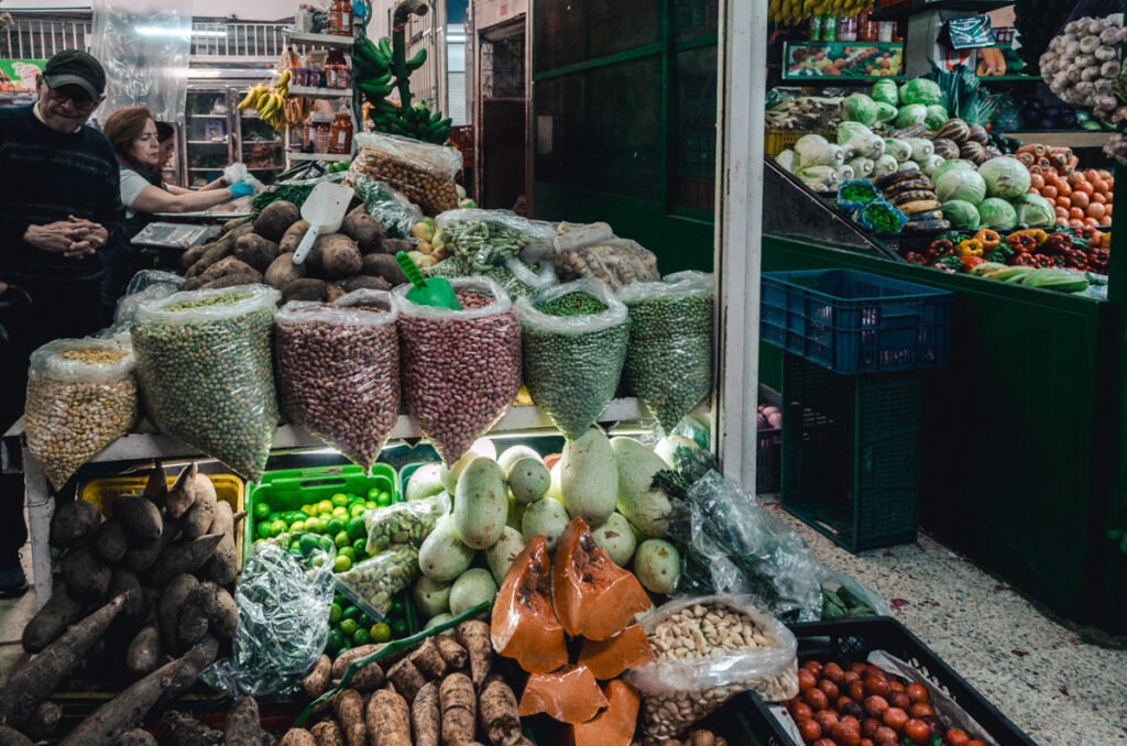 A variety of legumes and tubers neatly displayed in a stall at Paloquemao market, Bogota, Colombia, with beans and lentils in hanging bags and fresh produce like yuca and bananas in the foreground.