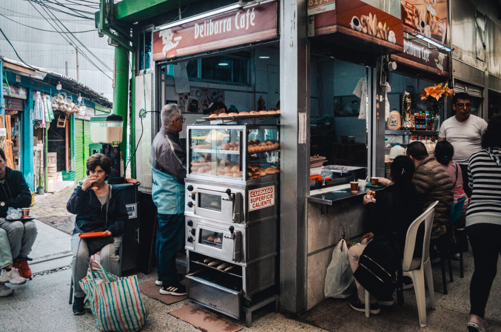Patrons enjoying fresh pastries and coffee at the Delibarra Café, a cozy corner stand in Paloquemao market, Bogota, with a sign warning 'Cuidado Superficie Caliente' (Careful, Hot Surface).
