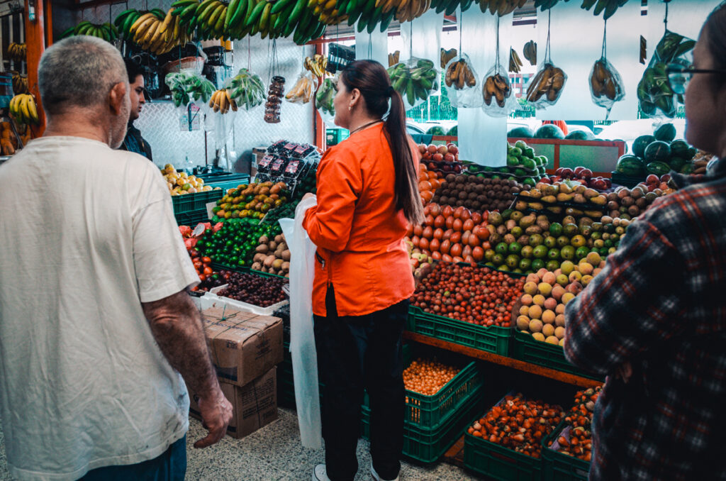 A merchant in an orange jacket attentively oversees a vast array of colorful fruits, including tomatoes and citrus, at a bustling stall in Paloquemao market, Bogota.