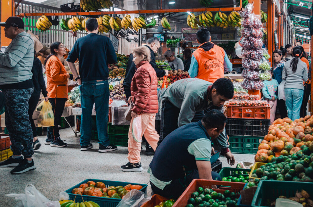 A lively scene at Paloquemao market, Bogota, with vendors and customers engaged in the vibrant trade of tropical fruits, vegetables, and hanging bananas.