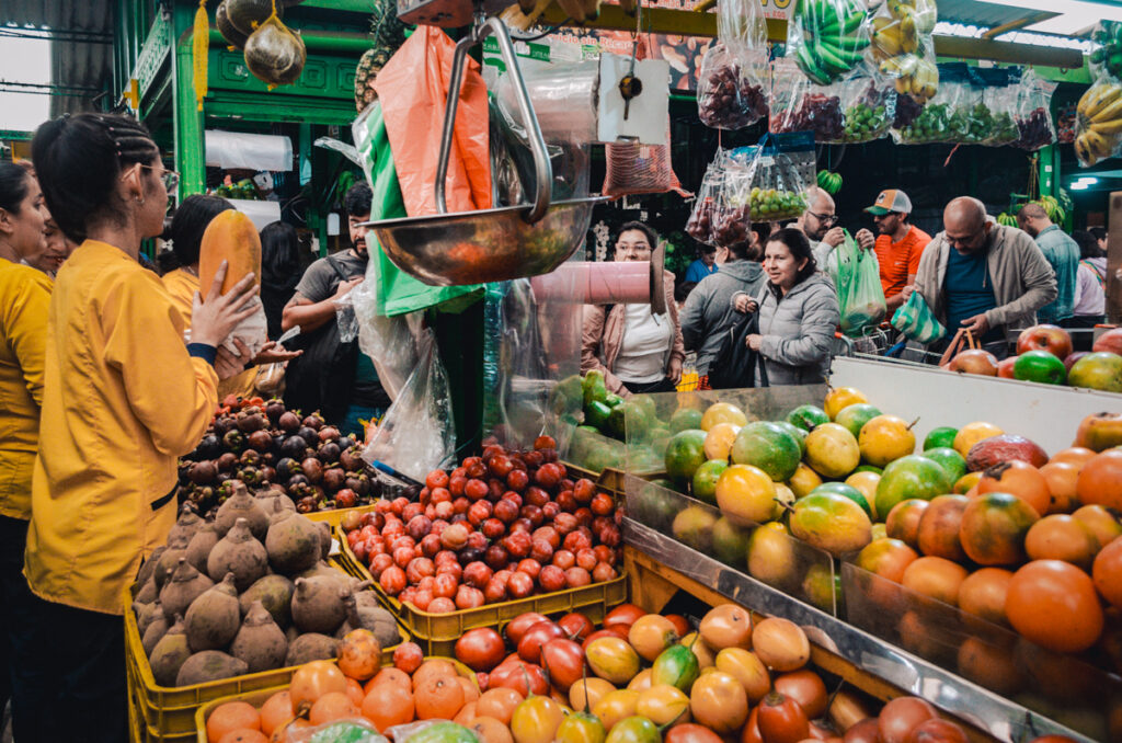 Busy scene at Paloquemao market in Bogota, Colombia, with customers engaging with vendors amidst a colorful array of fruits including mangos, oranges, and apples, and a woman in yellow examining a large papaya.