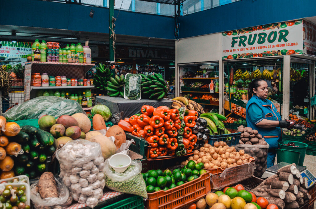 Vibrant display of fresh produce at Paloquemao market, including tomatoes, bananas, and various tropical fruits, with a vendor in a blue apron sorting through the items.