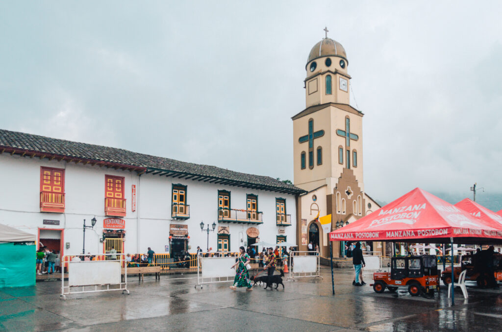 Plaza de Bolívar in Salento, Colombia, on a rainy day with the iconic yellow and white church standing tall against a cloudy sky. Vendors under red branded tents and locals with umbrellas add life to this vibrant town square.