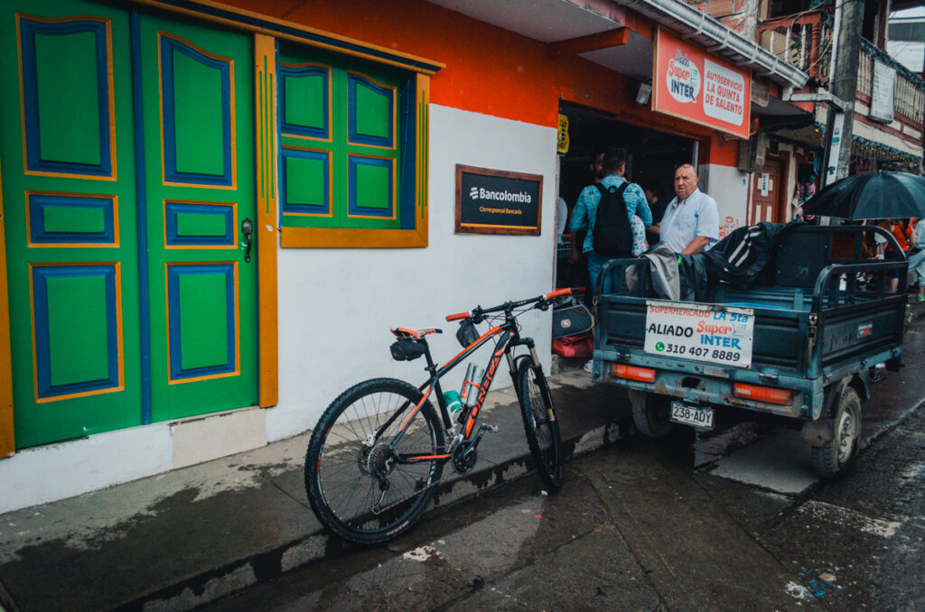 A mountain bike leans against the colorful facade of a building in Salento, Colombia, while daily life unfolds with locals interacting near a Bancolombia service point and a delivery vehicle marked 'Supermercado La 5ta'.