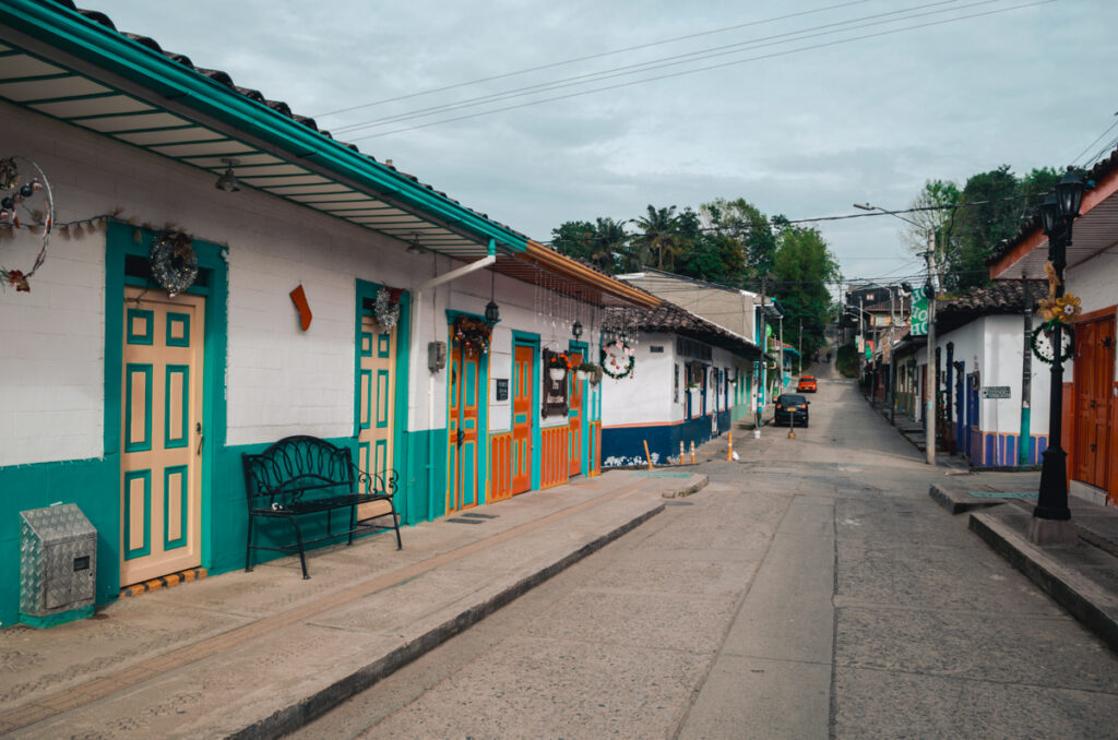 A tranquil street in Salento, Colombia, showcasing the charming colonial architecture with brightly painted doors and traditional red-tiled roofs. Streetlamps and festive wreaths add to the quaint atmosphere of this coffee region town, with a backdrop of lush greenery under an overcast sky.
