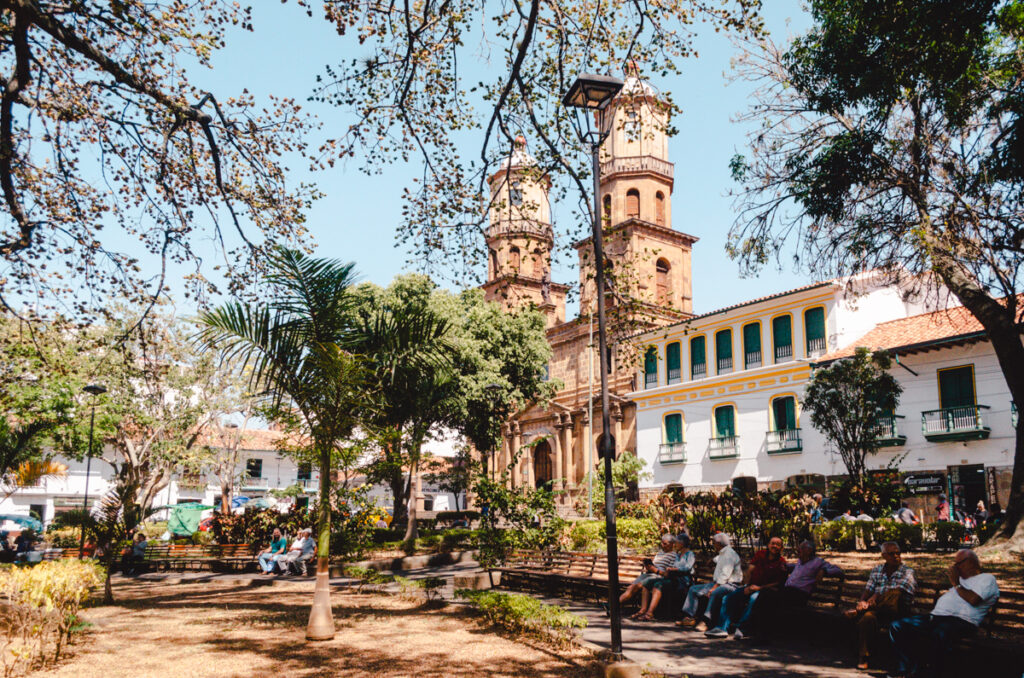 Bustling central park in San Gil, Colombia, with residents enjoying the shaded benches and the historic Church of San Gil standing proudly in the background, amidst tropical foliage under a sunny sky.