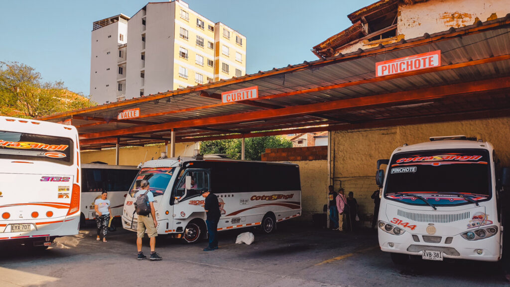 The image depicts the bustling bus terminal 'Terminalito' in San Gil , Colombia with multiple white buses, adorned with colorful graphics and labeled with destinations such as "VALLE," "CURITI," and "PINCHOTE." People are milling about, some boarding buses, while others appear to be waiting or conversing. The scene is set against a clear blue sky, with a multi-story building in the background and a weathered structure to the right. This is a typical day at a Latin American bus station, capturing the liveliness of local transport.