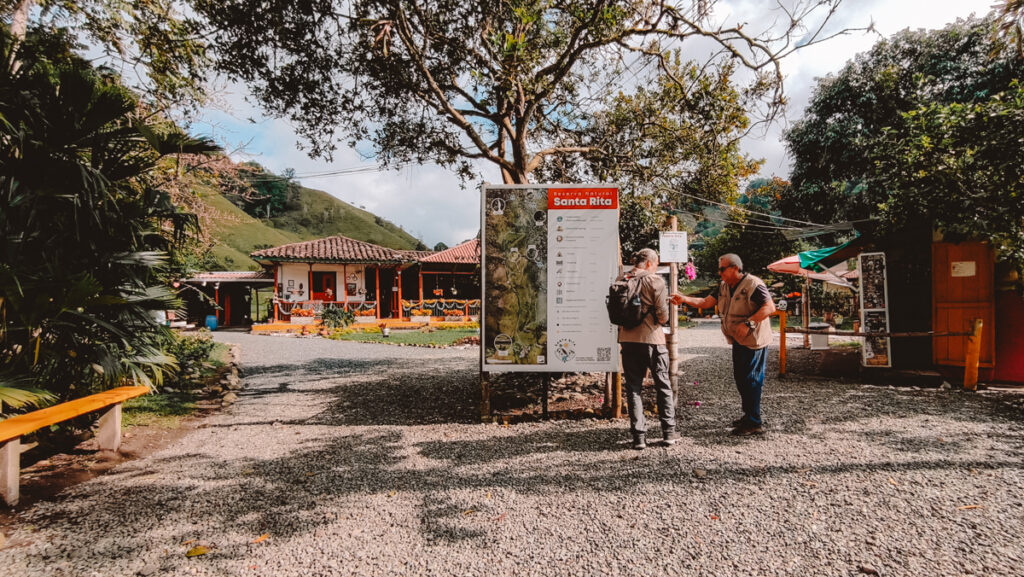 Visitors gather at the starting point of the Santa Rita nature trail near Salento, examining a large information board, with a quaint Colombian finca and lush landscape in the background, under a clear blue sky.