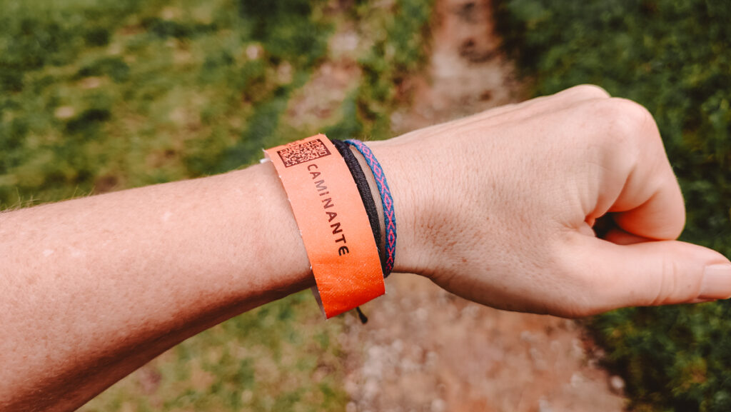 Person's wrist with a vibrant orange event wristband featuring the word 'CAMINANTE' and a QR code, alongside a blue and red friendship bracelet, with a blurred green background hinting at an outdoor setting. This wristband is a memento from the Santa Rita Salento adventure.