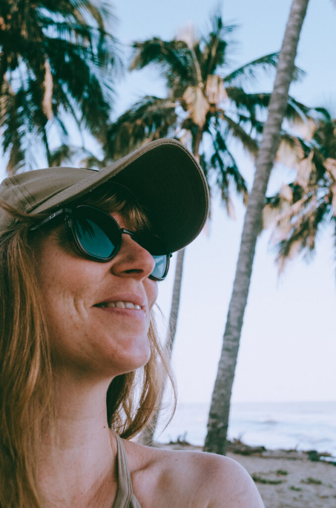 Smiling woman with shoulder-length hair wearing Sunski sunglasses and a cap, enjoying a tropical beach setting with palm trees in the background.