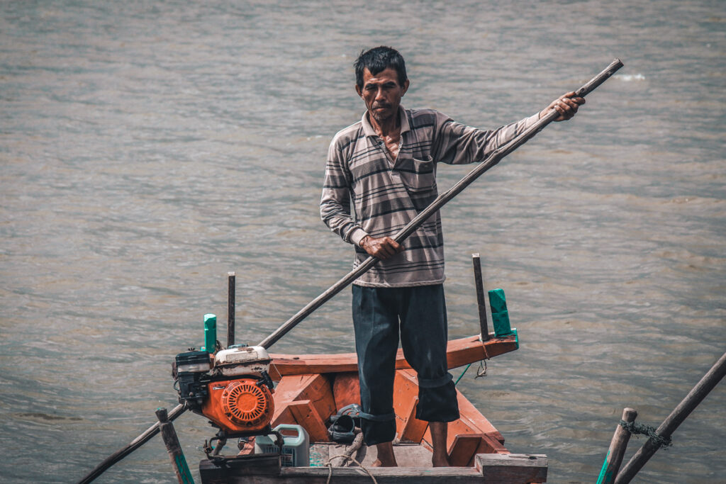 A man navigating a wooden boat with a long pole on the calm waters of Trapeang Sangkae in Kampot, Cambodia, with greenery along the riverbank.