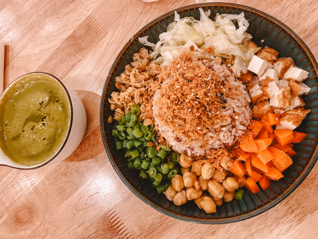A top view of a delicious vegan meal in Kuala Lumpur consisting of a plate with brown rice topped with fried shallots, diced tofu, chopped scallions, cooked chickpeas, steamed carrots, and sliced cabbage, served with a side of vibrant green smoothie in a white cup.