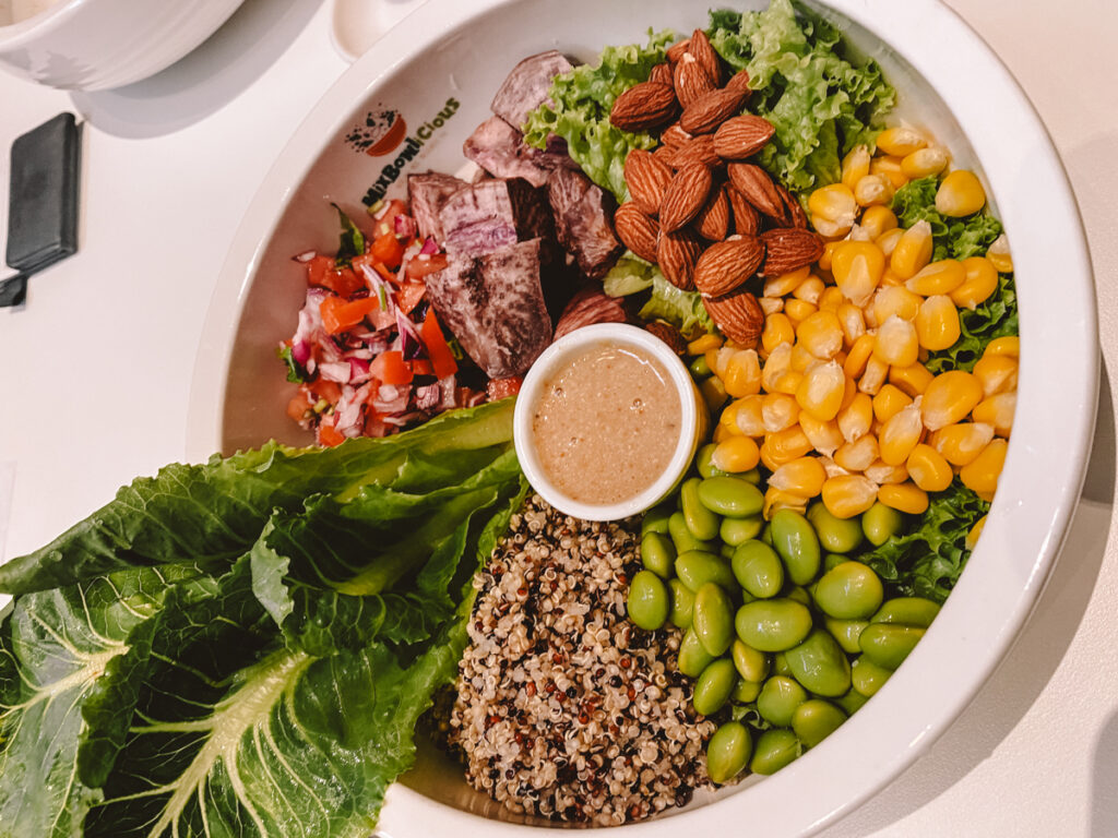 A healthy vegan food platter in Kuala Lumpur showing a balanced meal with lettuce leaves, diced tomatoes, red onions, gourd seeds, green soybeans, tri-color quinoa, and almonds, accompanied by a sesame dressing.