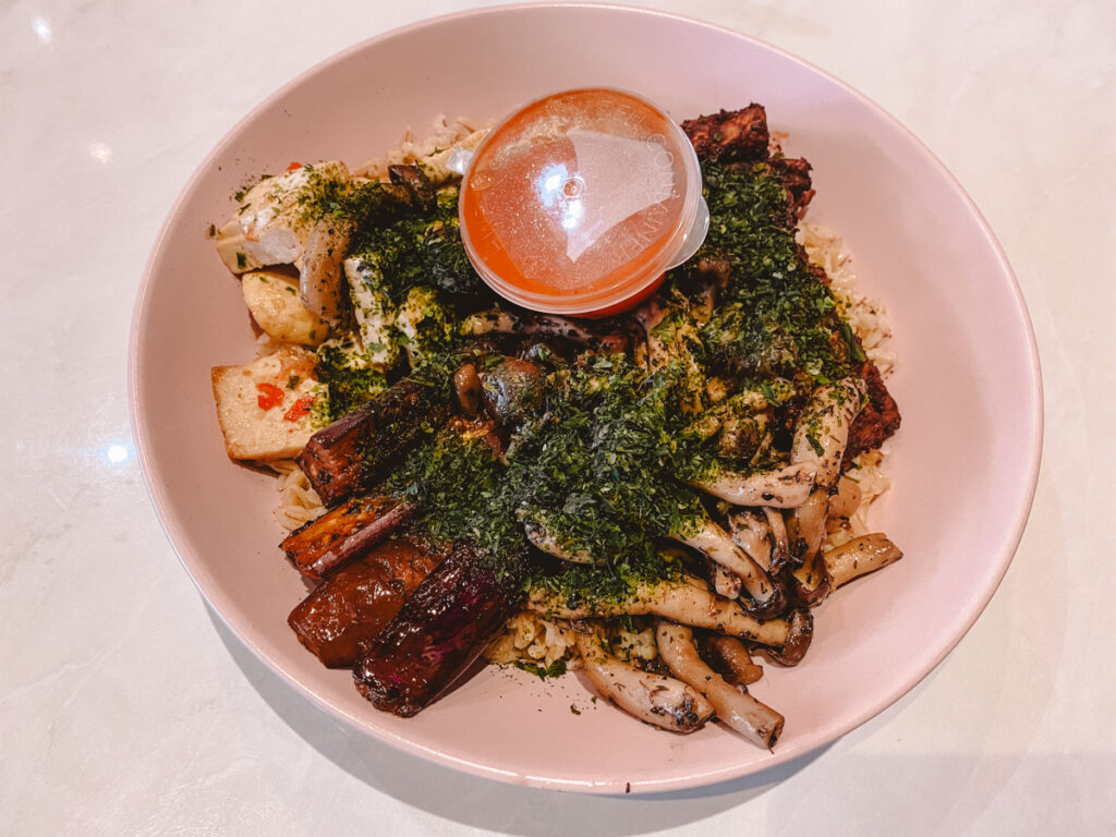A top view of a vegan dish in Kuala Lumpur featuring a bed of rice with roasted mushrooms, eggplant, and potatoes, topped with a generous sprinkle of chopped herbs and served with a side of tangy sauce.