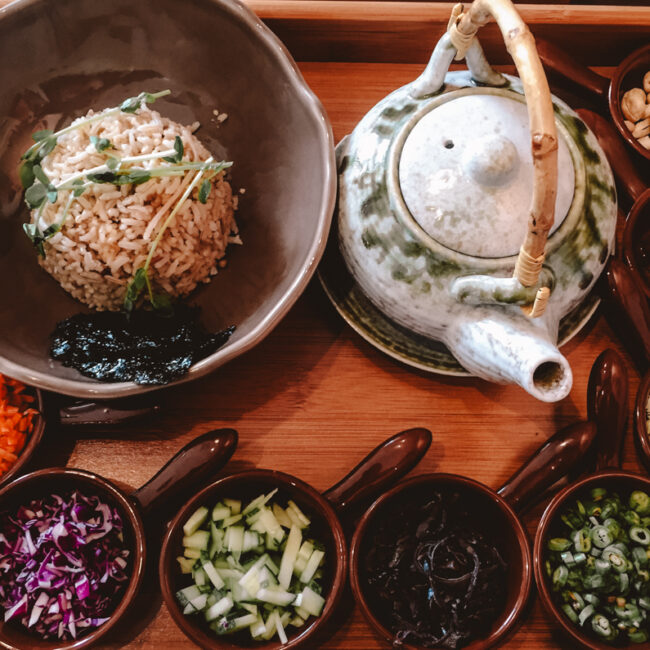 A traditional vegan food set in Kuala Lumpur displayed on a wooden tray, including a bowl of rice with greens, surrounded by small bowls of diced vegetables, seaweed, nuts, and pickled items, with a teapot on the side.