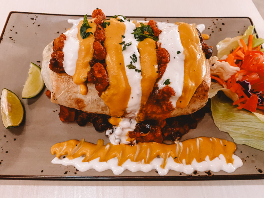 A vibrant plate of vegan food in Kuala Lumpur showcasing a stuffed bread roll topped with chili, vegan cheese, and a drizzle of creamy sauce, served with lime wedges and a side salad of lettuce, tomato, and grated carrots.