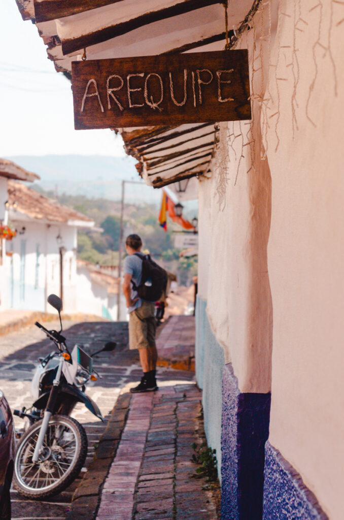 A quaint street in Barichara, Colombia, with a rustic wooden sign reading 'AREQUIPE' hanging from a whitewashed building. A man with a backpack pauses to admire the view, next to a parked motorcycle, under the shade of overhanging roof tiles.