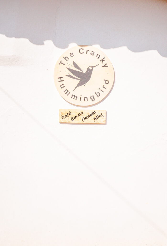 Simple yet charming sign of 'The Cranky Hummingbird' cafe in Barichara, Colombia, displayed on a white wall. The circular wooden sign features a stylized black hummingbird, complemented by a smaller sign listing offerings such as coffee, cacao, and honey.