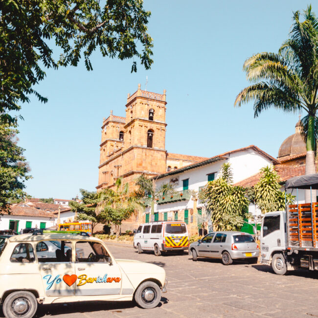 The vibrant main square of Barichara, Colombia, featuring the town's iconic brick church with ornate twin towers. Lush palm trees, a traditional tuk-tuk, and a vintage car adorned with 'Yo ❤️ Barichara' enhance the local charm under a clear blue sky.