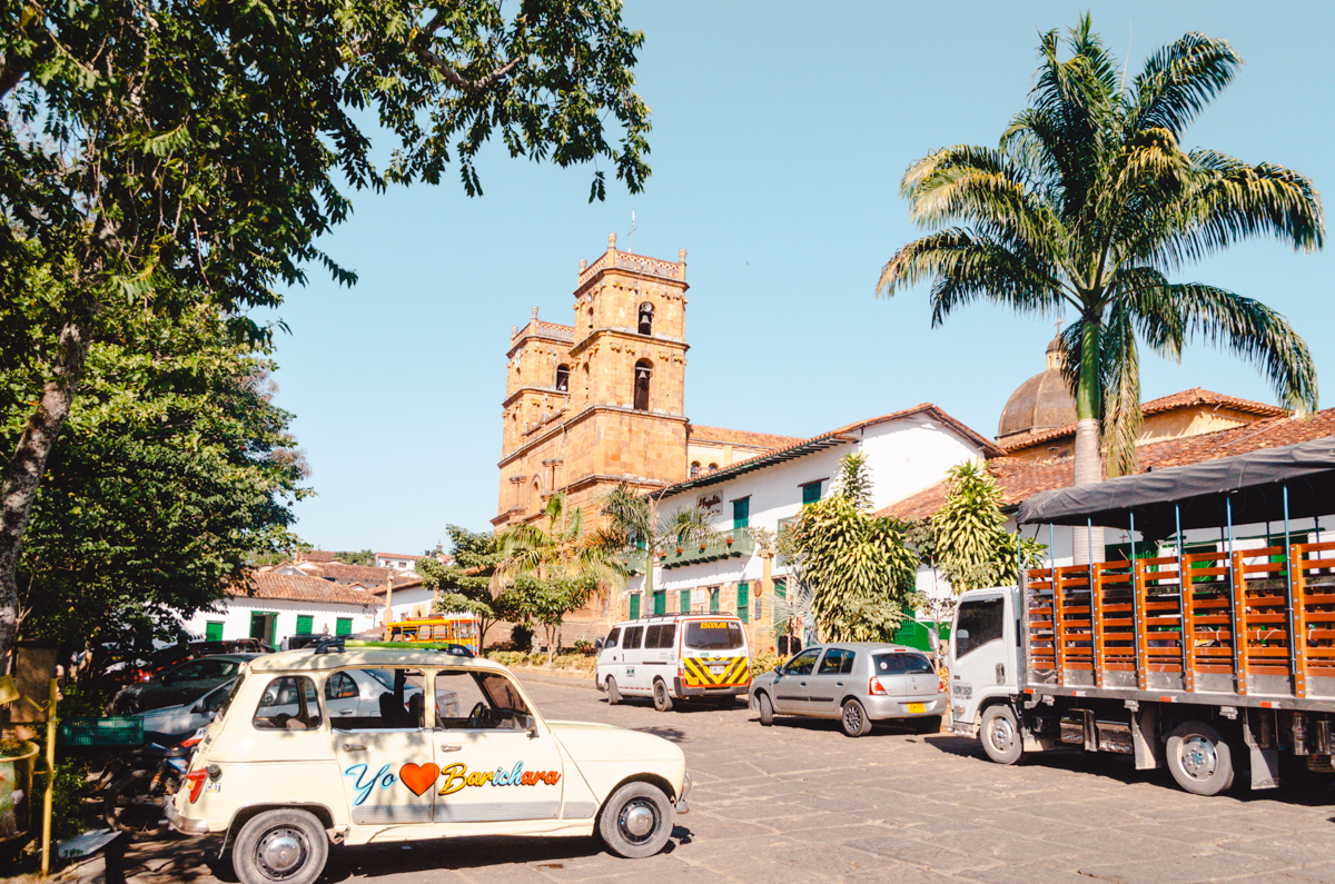 The vibrant main square of Barichara, Colombia, featuring the town's iconic brick church with ornate twin towers. Lush palm trees, a traditional tuk-tuk, and a vintage car adorned with 'Yo ❤️ Barichara' enhance the local charm under a clear blue sky.