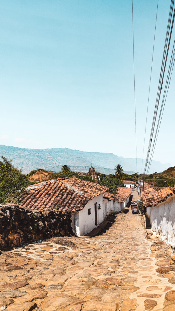 Historic stone-paved street in a colonial village called Guane in Colombia, leading downhill between old, white-walled houses with traditional clay roofs. The street, flanked by overhead power lines, provides a picturesque view of the lush countryside and distant mountains, embodying the charm of rural Colombia.