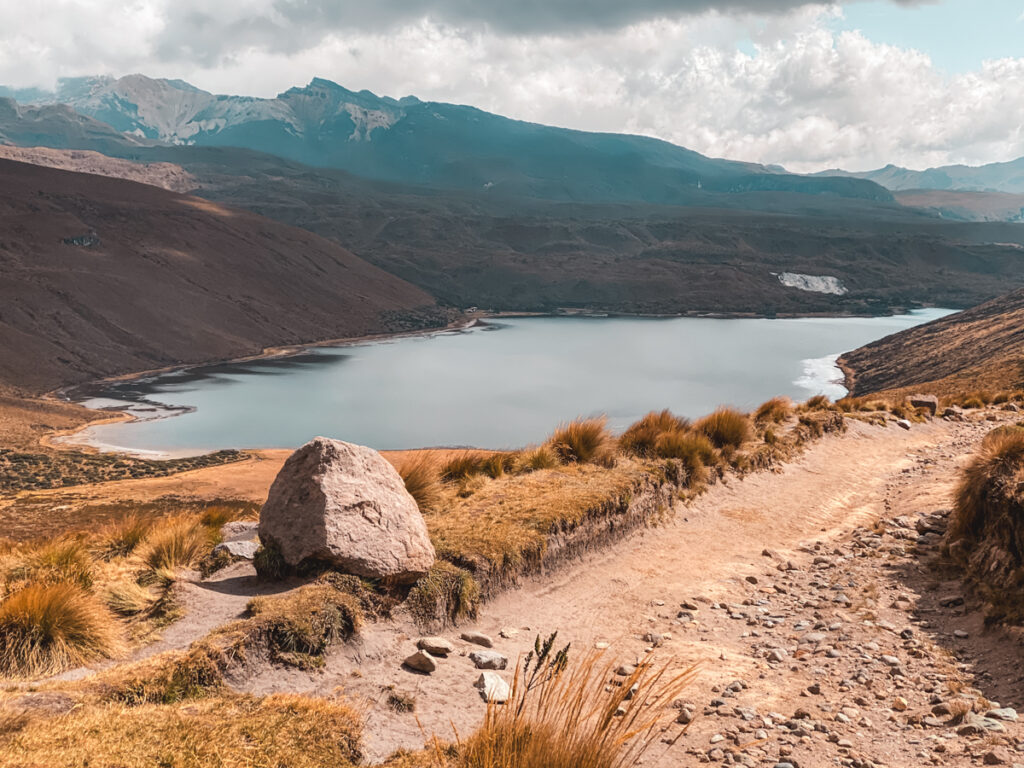 This image depicts the serene Laguna Otún, located in Los Nevados National Park, Colombia. The scene is framed by a rocky path leading towards the tranquil blue lake, surrounded by golden grasses and distant mountains, under a partially cloudy sky, creating a peaceful natural setting.