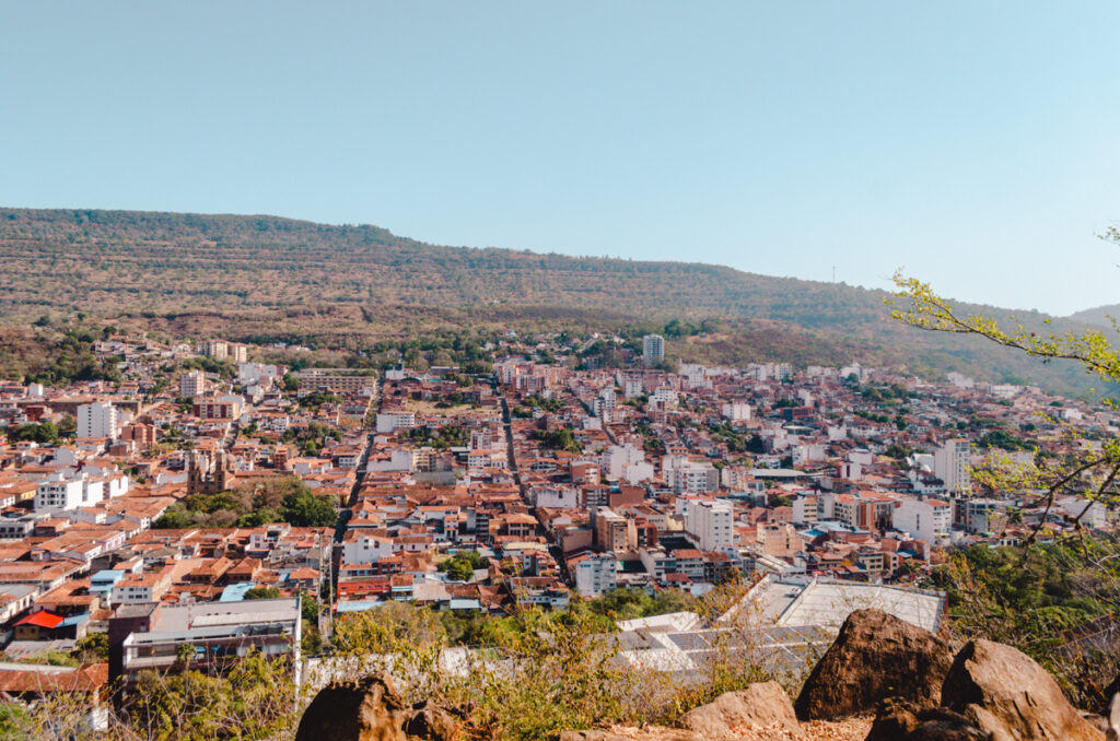 Elevated view from the Cerro de la Cruz viewpoint overlooking San Gil, Colombia. The landscape shows a dense tapestry of red-roofed houses, multiple winding streets, and modern buildings sprawling into the forest-covered hills under a clear blue sky.