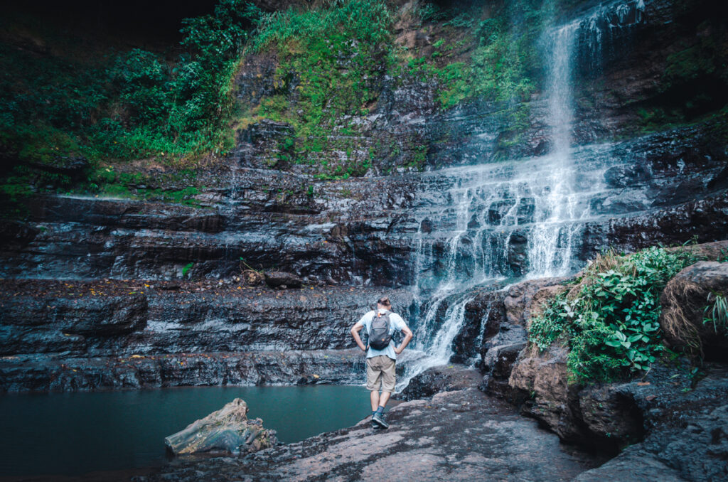 A man standing in front of the majestic Juan Curi waterfall, captivated by its beauty and the soothing sound of rushing water.