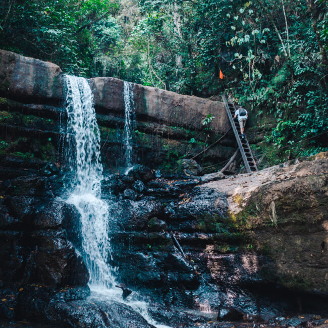 A smaller waterfall at Juan Curi in San Gil, where water spills over a broad, flat rock into a serene pool. A person climbs a ladder on the rock face, adding a sense of scale to the lush, forested environment.