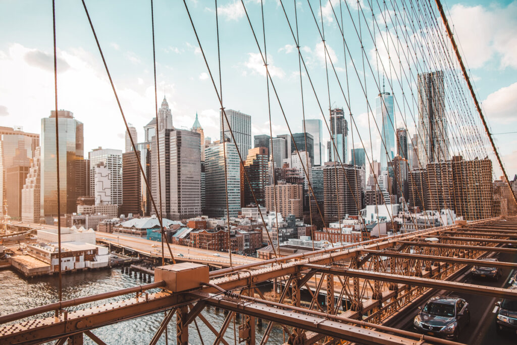 This is a photo taken from the top of Brooklyn Bridge. You can see cars driving over the bridge below and there's a panoramic view over the city in the background on a sunny morning.