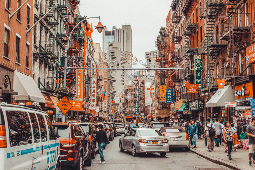 This is a photo of a busy street in Chinatown. the red brick buildings all have Chinese signage on their walls, there's a grey car in the middle of the street and a lot of pedestrians on the sidewalk.