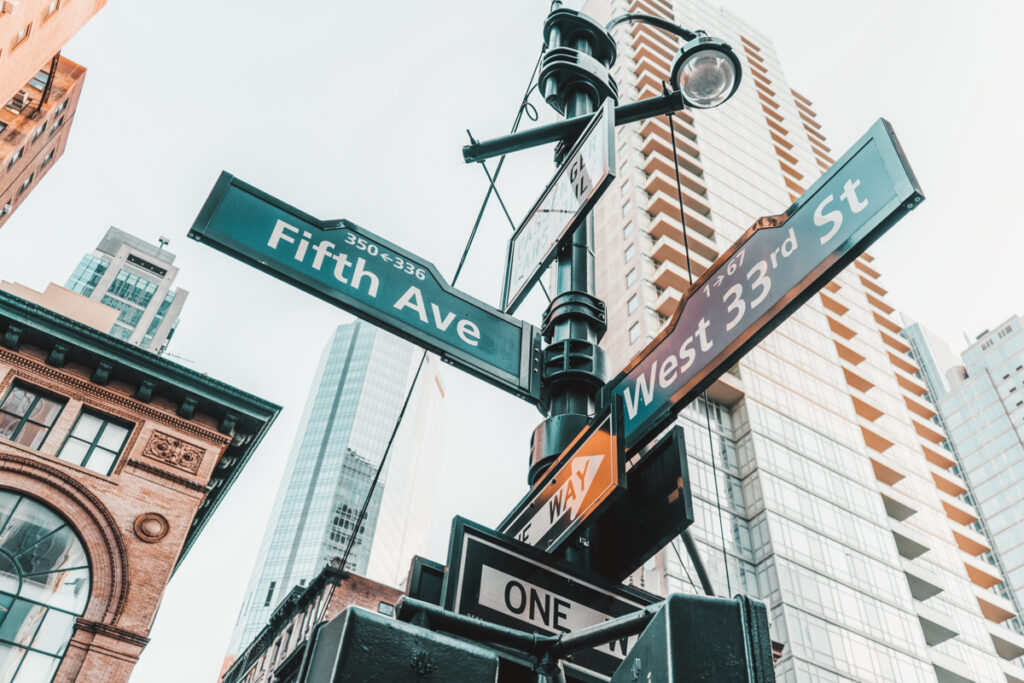 This photo shows street signage of fifth Avenue in New york. The photo is taken from the bottom of a lighting pole and you can see high-rise buildings in the background.