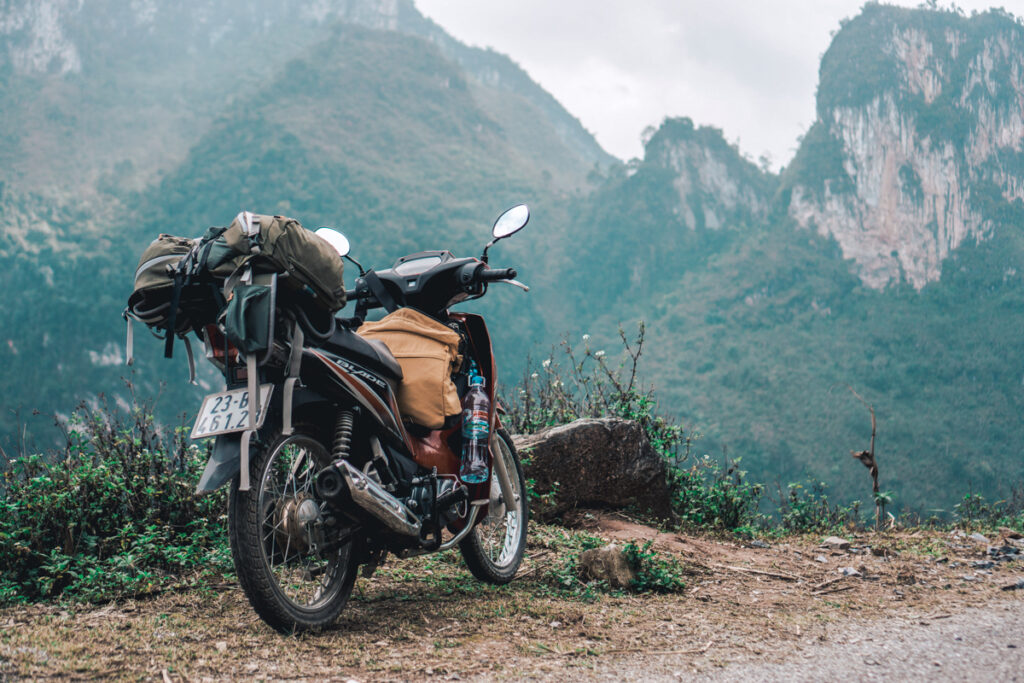 A motorbike standing on the side of a road in the Ha Giang region. There are mountains in the background and the motorbike is loaded with luggage.