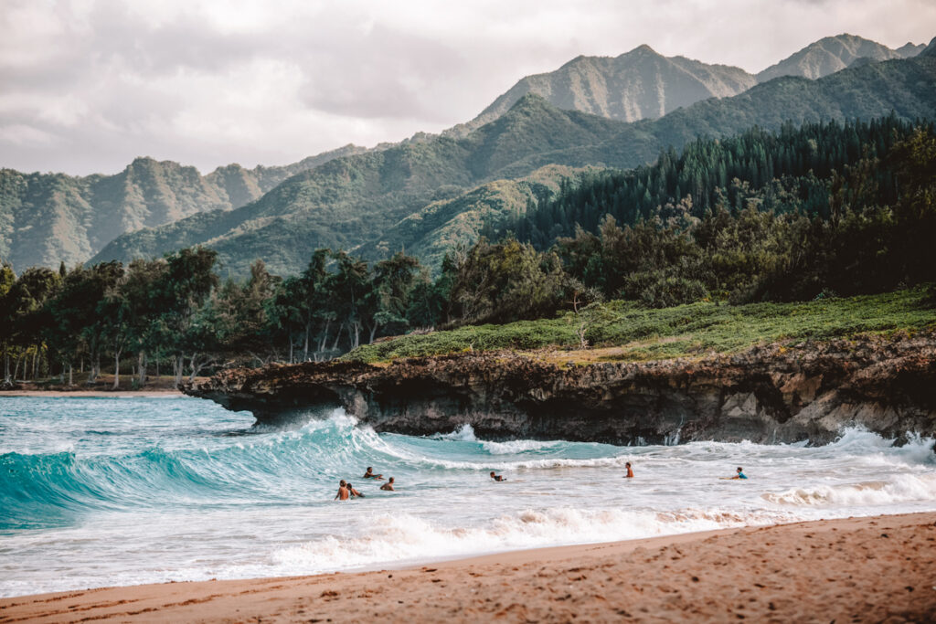 In this Hawaii puns photo, you can see a dark beige-colored beach and blue ocean with rolling waves. There are a few surfers in the water and in the background are lush green mountains.