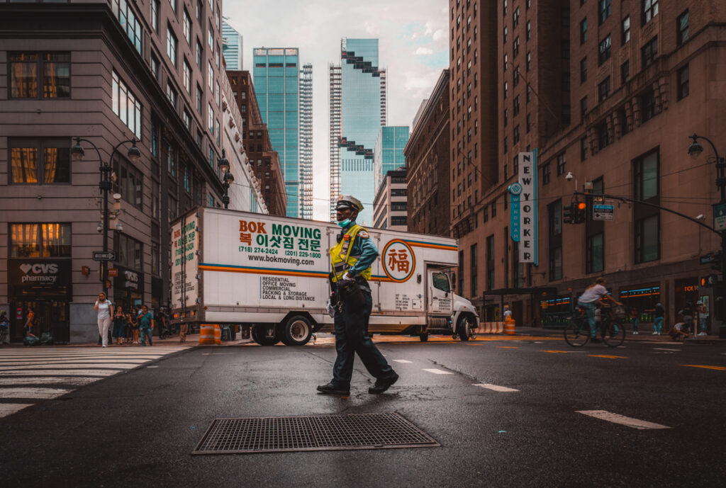 This photo shows a street in New York City. A police man is standing on a crossroads in between the high-rise buildings of the city. In the back is a white truck with signage on the side saying 'Bok Moving'.