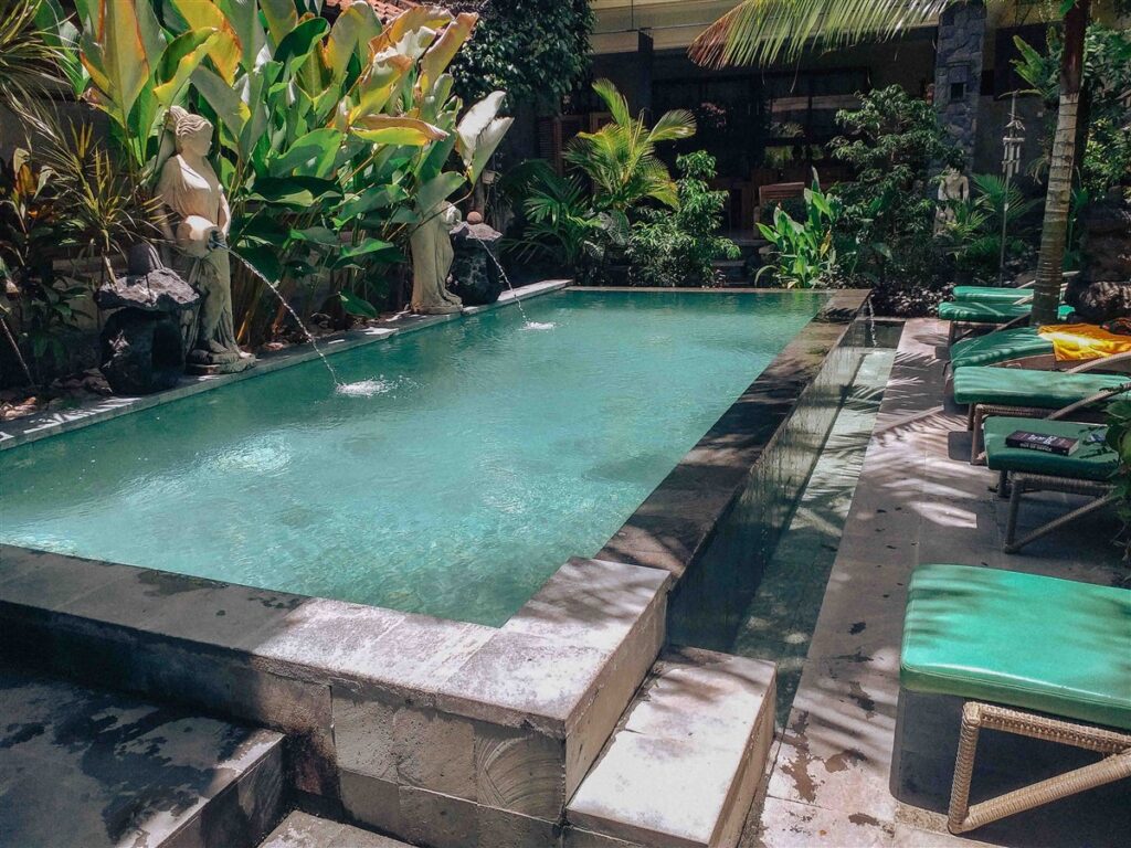 This is a photo of a beautiful square swimming pool in a small guesthouse in Sanur in Bali. It is made from grey stones and is surrounded by lush greenery.