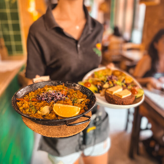 A lady presents two plates of food, one in the back is blurry but one in front presents a delicious vegan paella The interior of the restaurant is made from wood and hits of green.