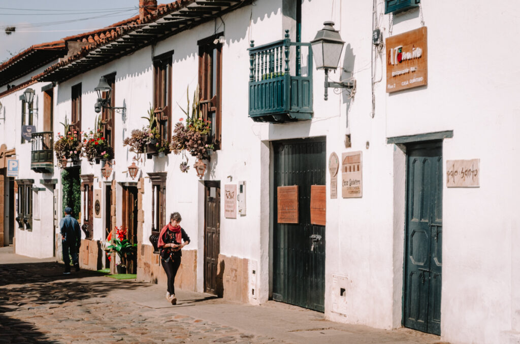 A woman walking in a a cute street in Villa de Leyva. The white houses have green doors and windows and are adorned with flowers.