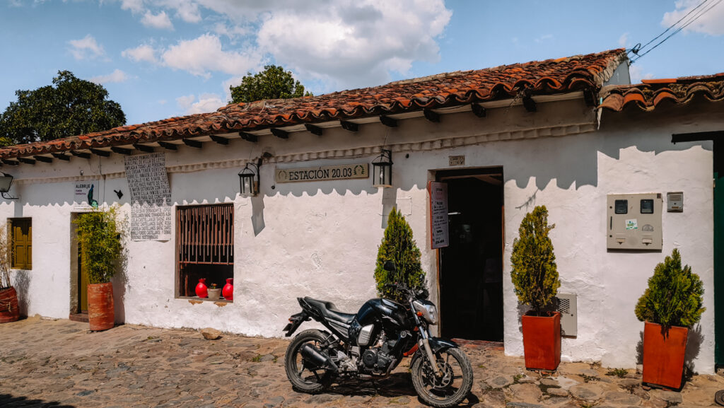 The white front of Estacion 20 03 in Villa de Leyva. There's a motorcycle in front of the door and red pottery with plants.
