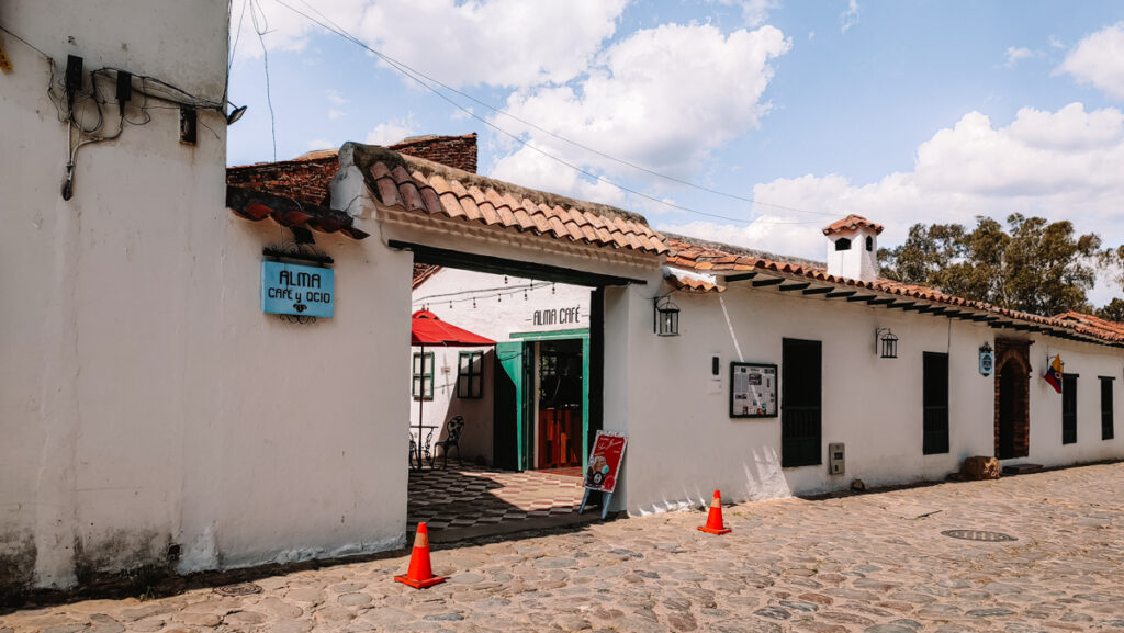 A white building with a gate leading to Alma Cafe in Villa de Leyva in Colombia. The sun is shining and there are a few clouds in the sky.