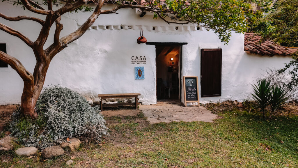 The facade of a white house in Villa de Leyva with a sign saying 'Casa Guadalupana'. There's a bit of grass in front of the door and a tree to the left of it.