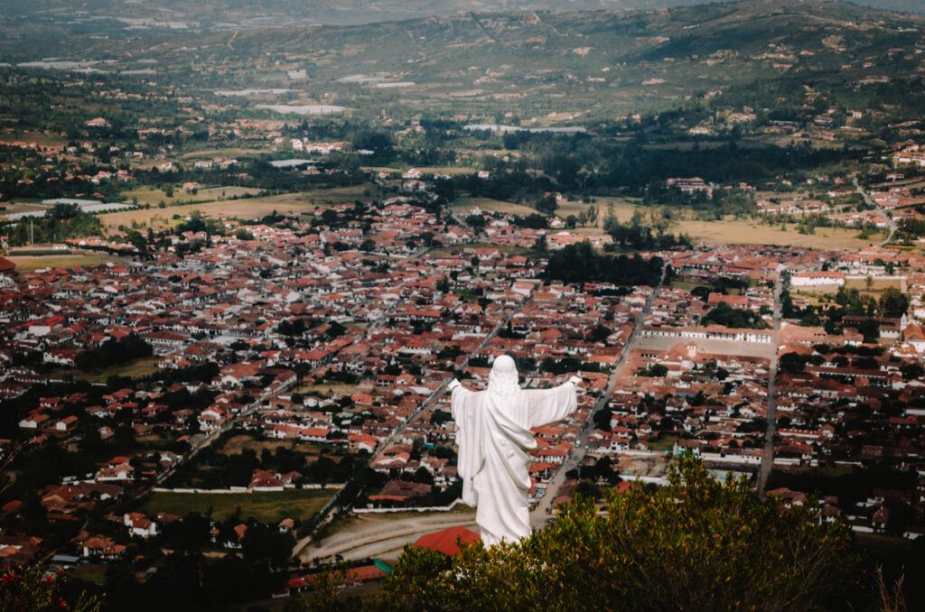 A panoramic view over Villa de Leyva from the top of a mountain. On the top of the mountain is a white Jesus statue looking down on the village.